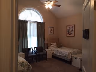 A small bedroom in an assisted living facility with two single beds, a nightstand with a lamp between them, two chairs near a large window with curtains, a ceiling fan with lights, and framed artwork on the walls.