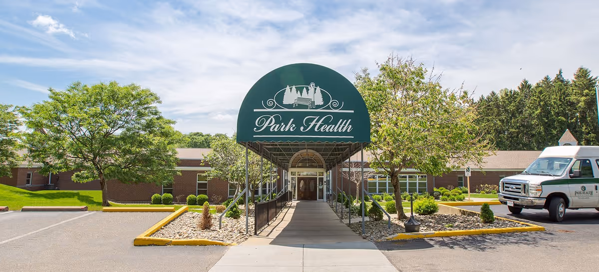 Entrance to Park Health Center with a green canopy displaying the facility's name. The building is a single-story brick structure surrounded by trees and landscaping. A white van with the Park Health logo is parked on the right side.