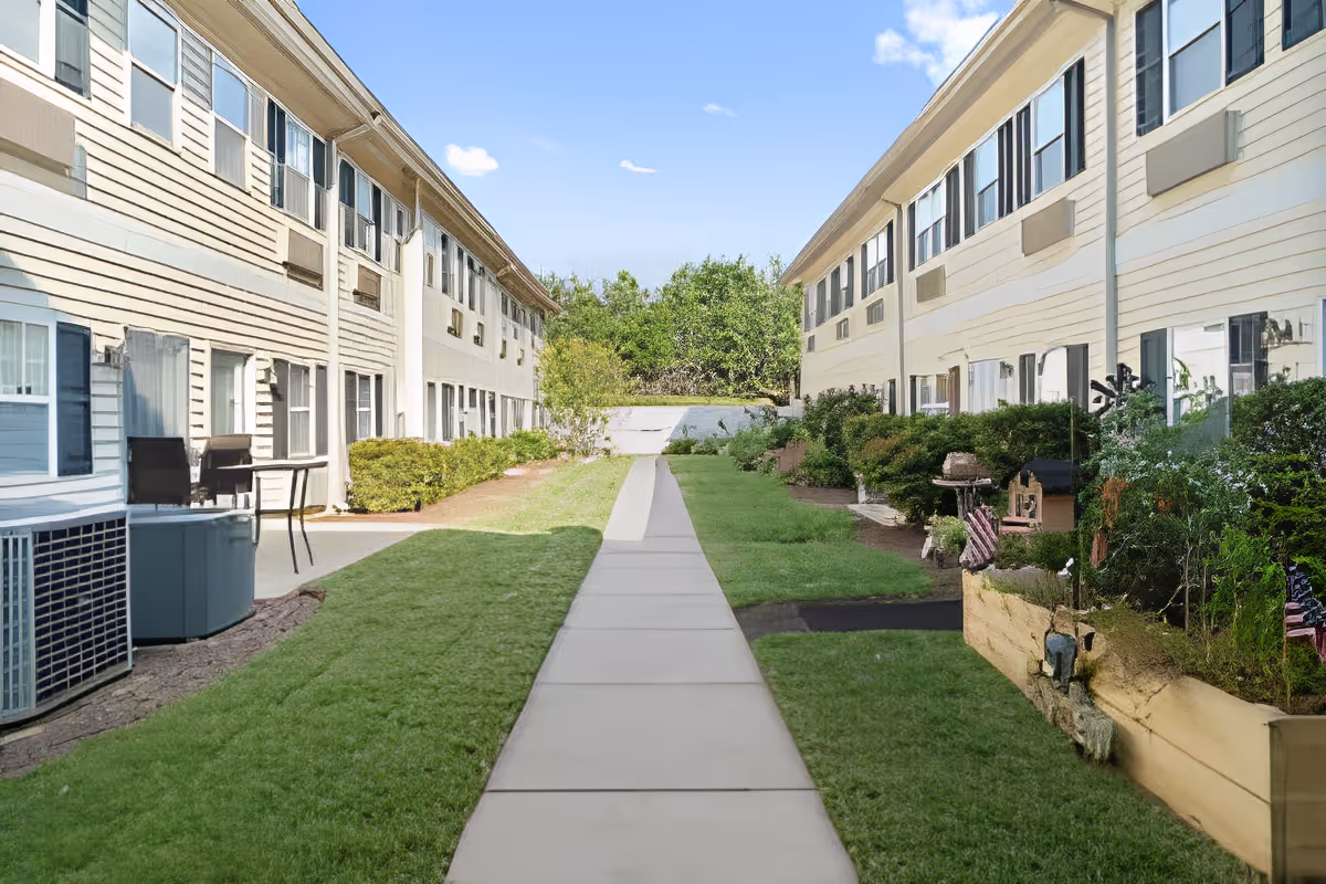 Outdoor view of a pathway between two beige two-story buildings with multiple windows. The pathway is surrounded by green grass, bushes, and small garden areas. The sky is clear with a few clouds.