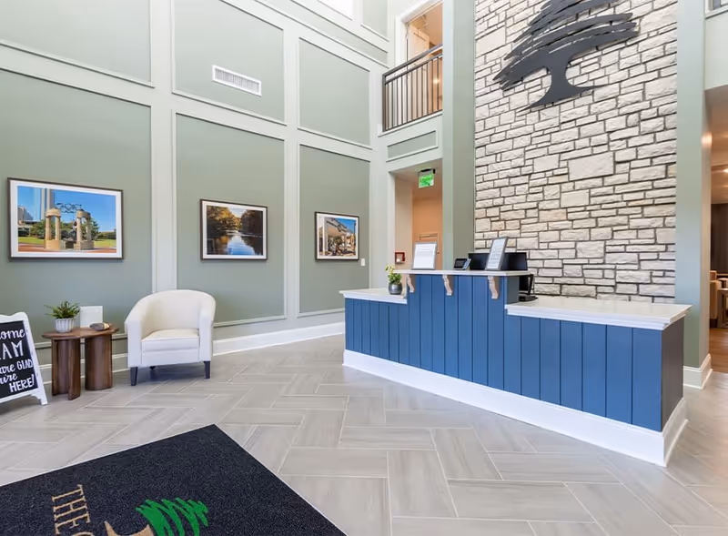 Reception area of Canopy at Westridge featuring a blue front desk with a white countertop, a stone accent wall with a large tree logo, light green walls with framed pictures, a white armchair, and a small wooden side table with a plant and a sign.