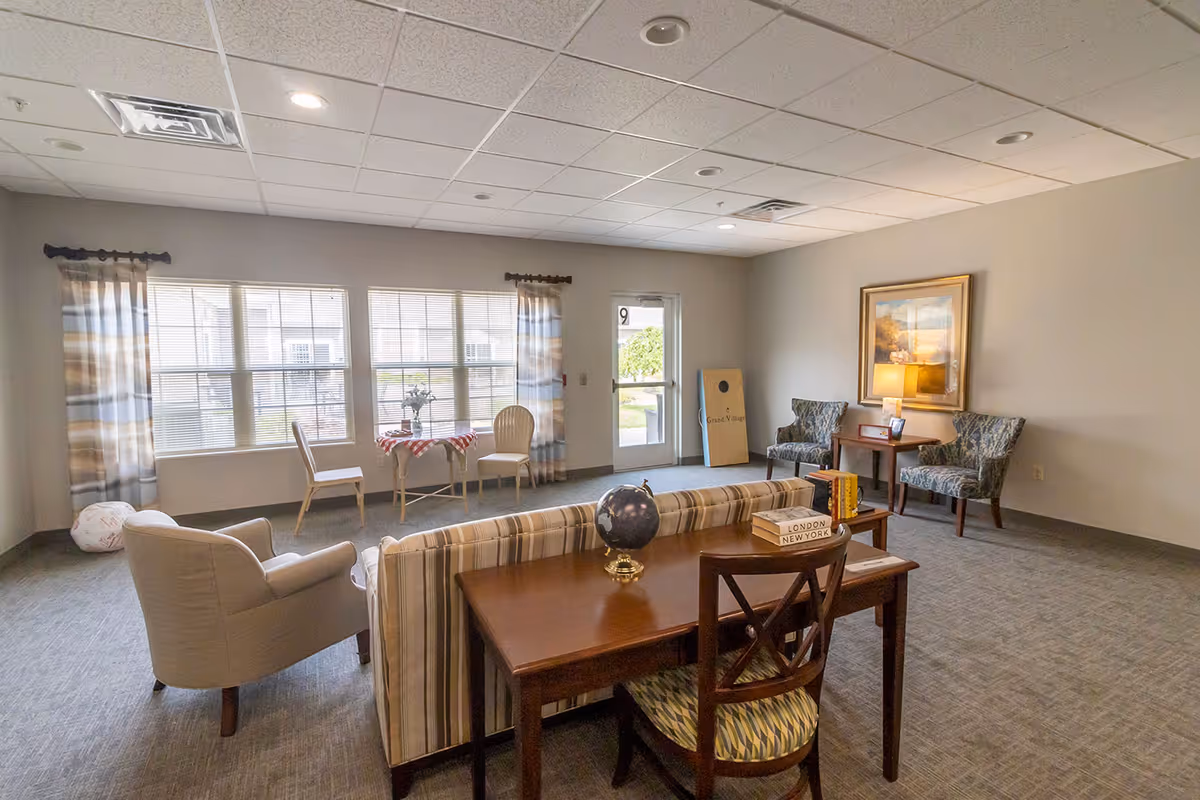 A spacious living room area in an assisted living facility with a striped sofa, a wooden desk with a globe and books, two patterned armchairs with a side table and lamp, a small table with two chairs near large windows with curtains, and a cornhole game board leaning against the wall near a door.