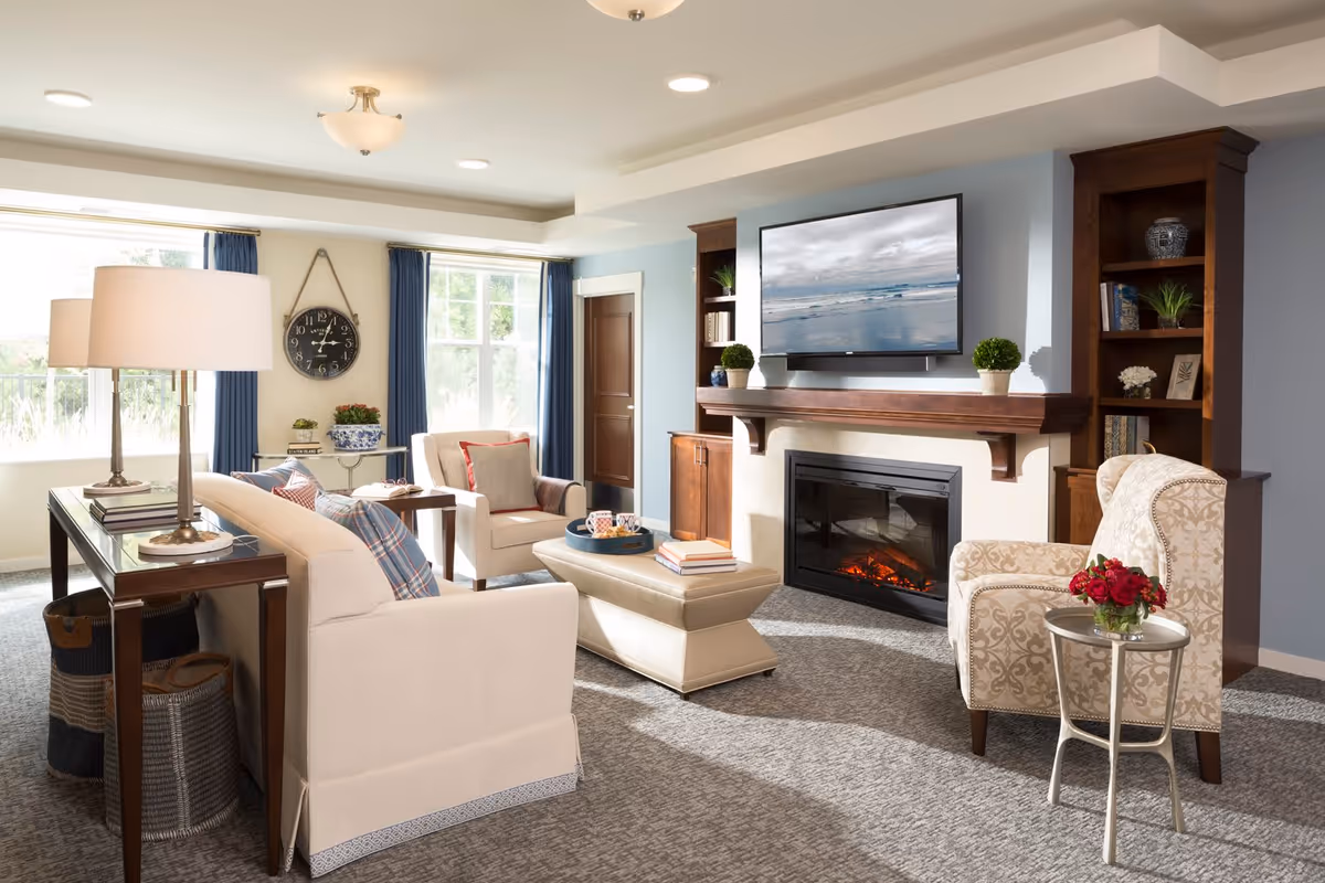 A cozy senior living room with beige and patterned armchairs, a beige sofa with plaid pillows, a rectangular ottoman, and a wooden console table with two lamps. The room features a fireplace with a wooden mantel, a flat-screen TV mounted above it, built-in wooden shelves with decorative items, large windows with blue curtains, and a round side table with a vase of red flowers.