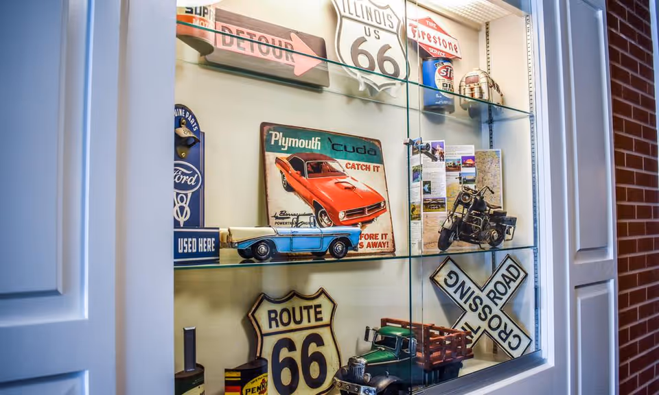 Glass display case with vintage automotive memorabilia including Route 66 signs, a Plymouth 'Cuda poster, model cars, a motorcycle model, and various retro signs and advertisements.