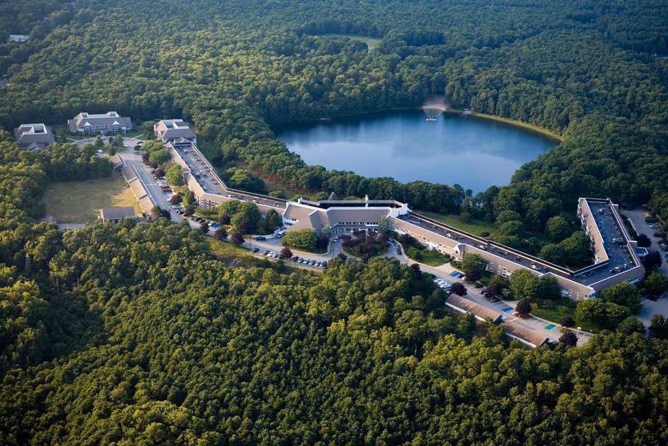 Aerial view of Thirwood Place, a large senior living facility surrounded by dense forest with a lake nearby. The building is long and sprawling with multiple wings and parking areas visible.