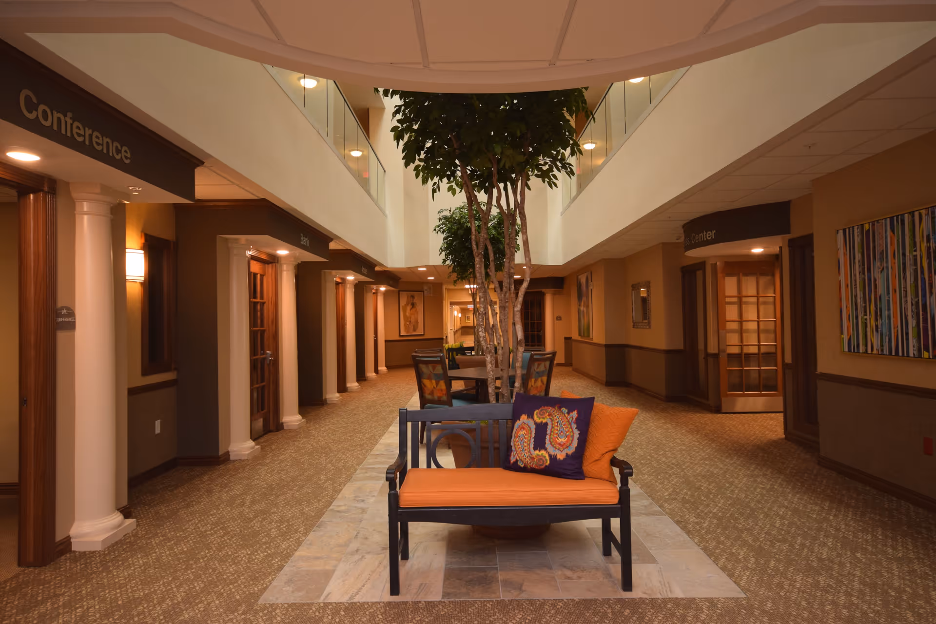 Interior hallway of Stone River Retirement Community featuring a central seating area with an orange cushioned bench and decorative pillows, a tall indoor tree, and multiple doors labeled Conference, Den, and Lanier. The hallway has warm lighting, carpeted floors, and artwork on the walls.
