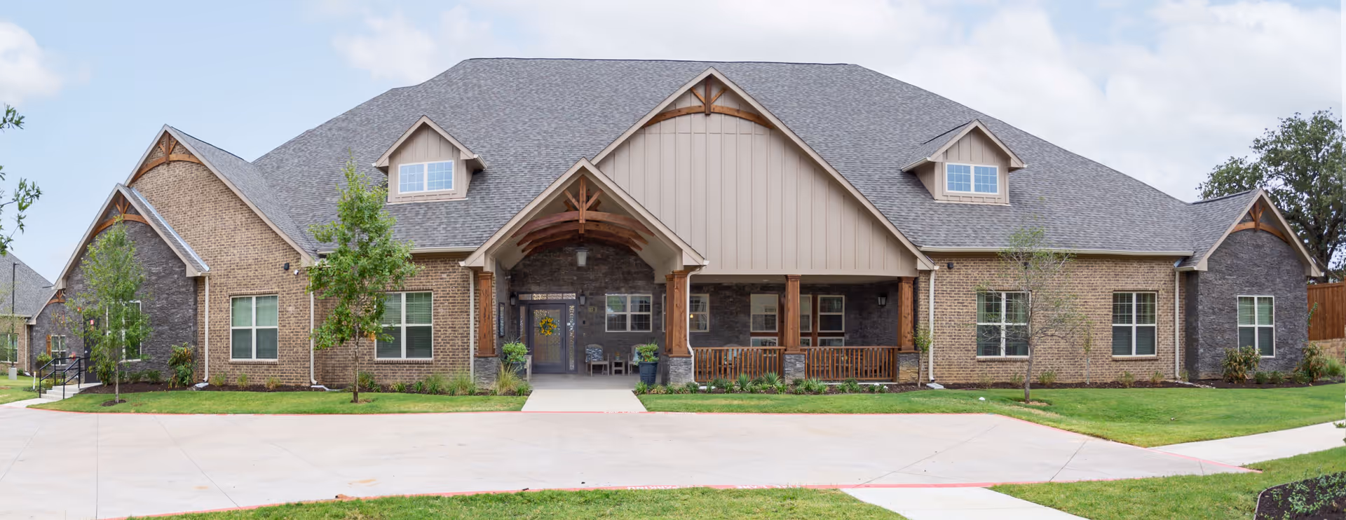 Front exterior of a long single-story brick building with a large gabled roof, central covered entrance with wooden posts and porch seating, and a paved driveway and lawn.