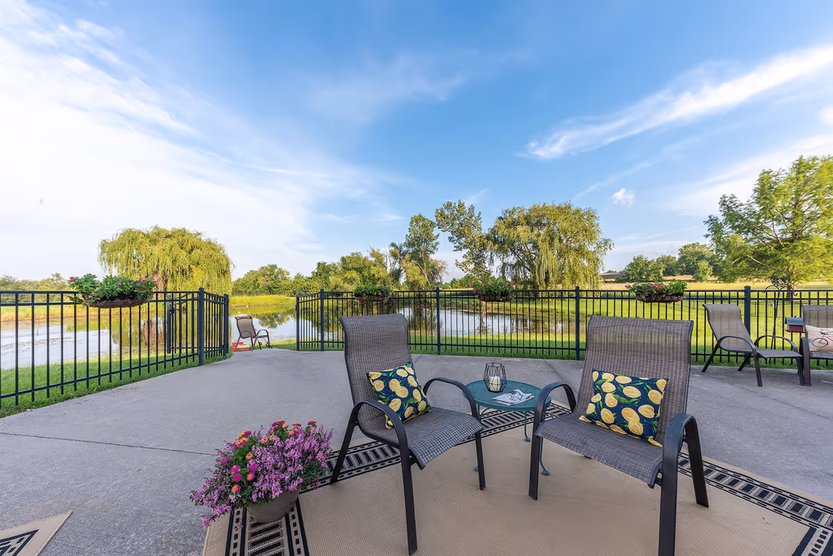 Outdoor patio area with several chairs and a small table on a concrete surface, overlooking a pond surrounded by green trees and grass under a blue sky with some clouds. Flower pots with blooming flowers are attached to the black metal fence enclosing the patio.