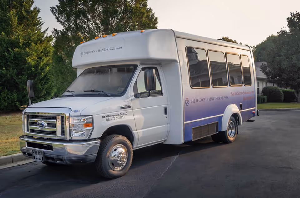 White shuttle bus with purple gradient parked on a paved lot beside trees.