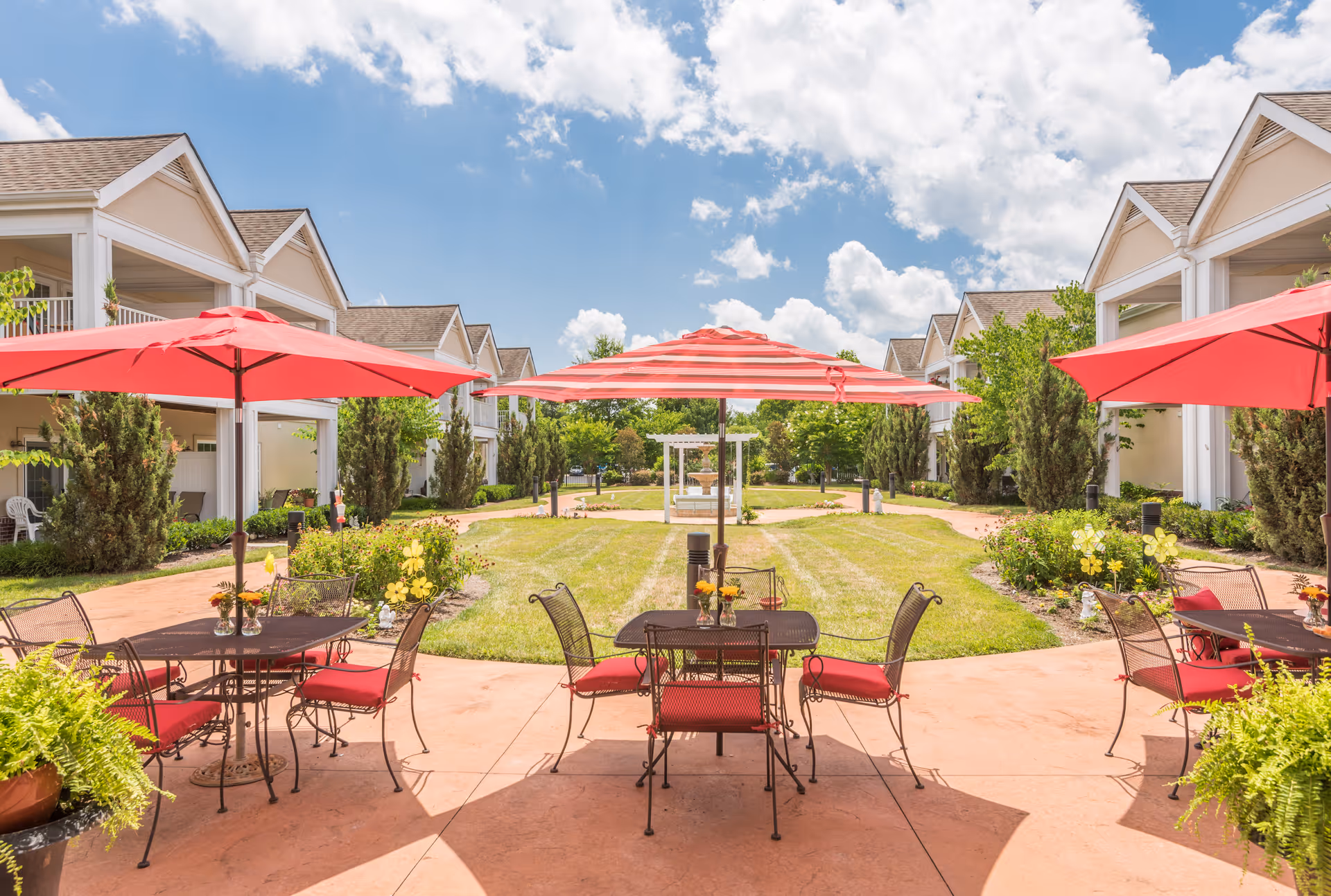 Courtyard patio with red umbrellas, metal tables and chairs between two rows of light-colored buildings under a blue sky.