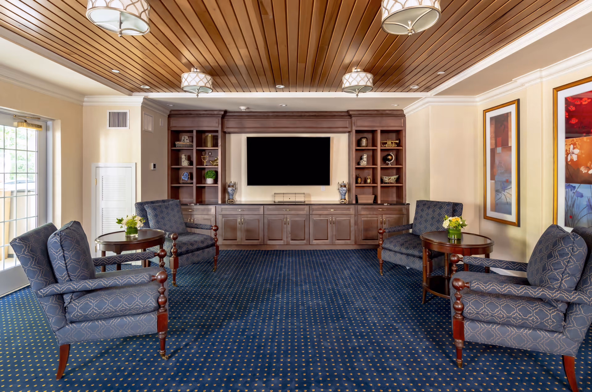 A cozy living room with four patterned blue armchairs arranged around two round wooden tables with small flower arrangements. The room features a blue carpet with a dotted pattern, a wooden ceiling with recessed lighting and hanging lamps, and a large built-in wooden entertainment center with shelves and a flat-screen TV. Two colorful framed artworks hang on the right wall, and a glass door on the left lets in natural light.