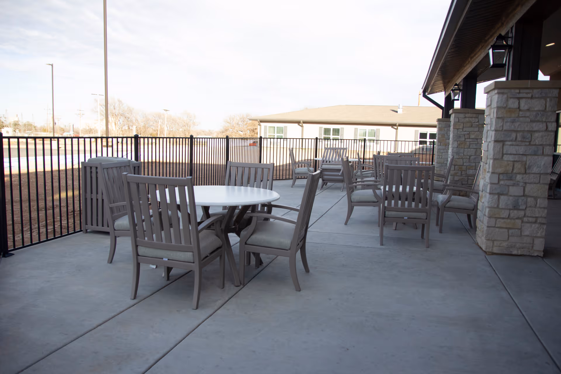 Covered outdoor patio with a round table, multiple cushioned chairs, stone pillars, and a metal fence.