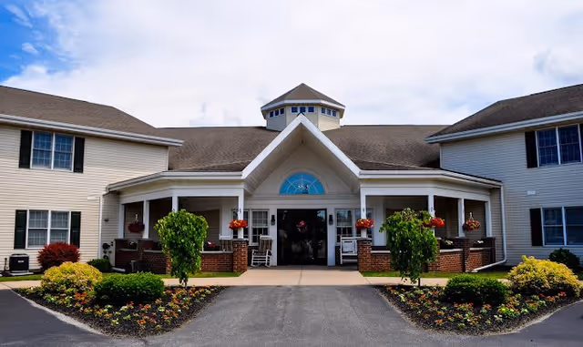 Front exterior view of a senior living facility named The Greens with a symmetrical building featuring a central entrance, white siding, brick accents, a peaked roof, and landscaped flower beds with small trees and shrubs along the walkway.