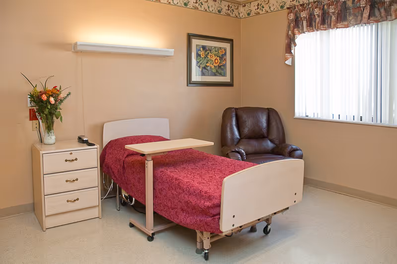 A simple bedroom in a senior living facility featuring a single hospital-style bed with a red patterned bedspread, an adjustable overbed table, a wooden nightstand with a vase of flowers and a telephone, a brown leather armchair, a window with vertical blinds and floral valance, and a framed floral painting on the wall.