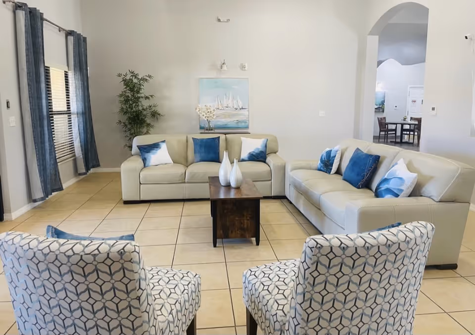 Bright communal living room with beige sofas, patterned chairs, blue-accent pillows, a central coffee table and wall art.