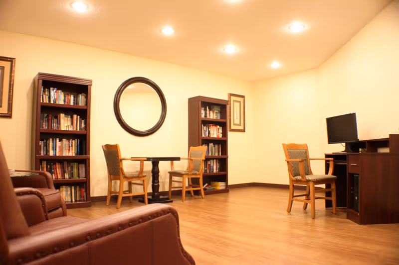 A warm common room with bookshelves, a round mirror, several chairs around a small table, and a computer desk.