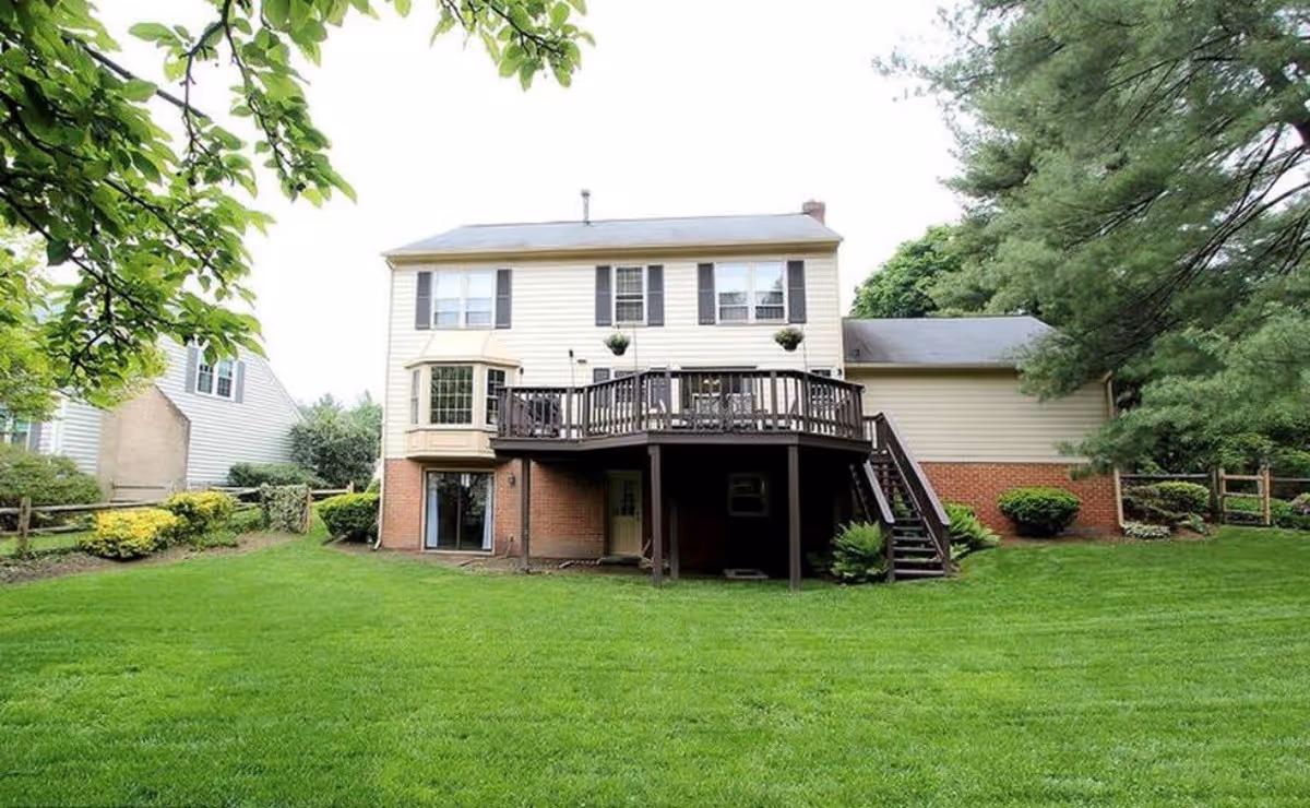 Rear exterior of a two-story house with a raised wooden deck and staircase overlooking a green lawn.
