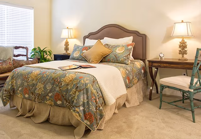 Sunlit furnished bedroom with a floral-patterned bed, two bedside tables with lamps, a chair and a window.