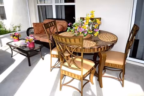 Outdoor patio area with a round table and four chairs, decorated with a floral centerpiece. Nearby, there is a cushioned bench and a coffee table with additional flower arrangements. The setting is bright and sunny, adjacent to a building with sliding glass doors.
