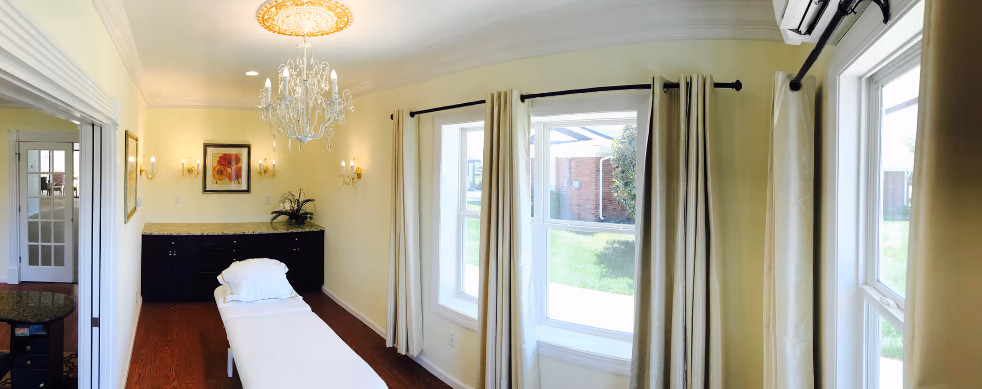 Bright interior room with a treatment table, chandelier, side cabinet and large windows with curtains.