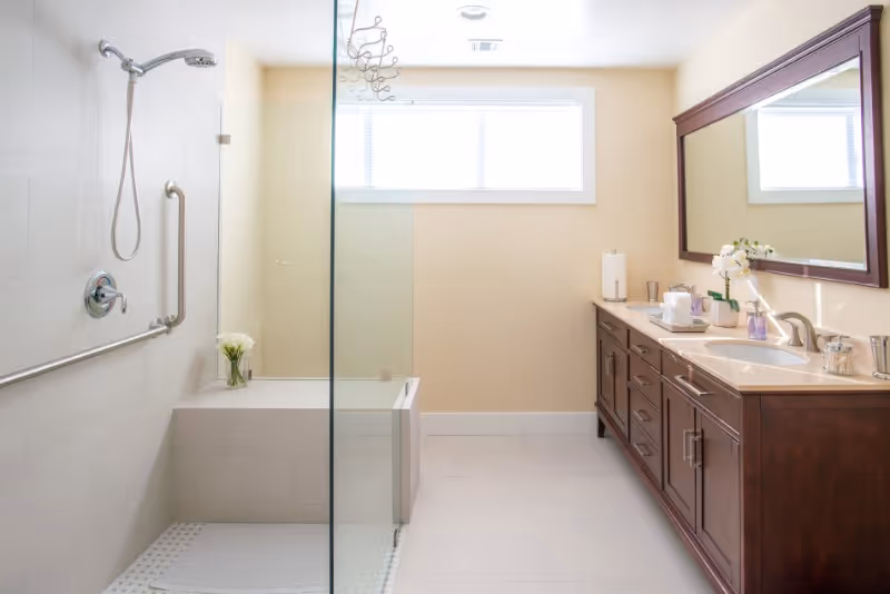 Bright and clean bathroom with a glass-enclosed shower area featuring a built-in bench, a handheld showerhead, and grab bars. Opposite the shower is a long wooden vanity with a large mirror, sink, and various toiletries. A window above lets in natural light.