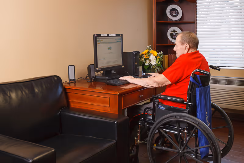 An elderly man in a wheelchair using a desktop computer on a wooden desk in a room with a black leather couch, a window with blinds, and a wooden shelf with decorative plates and books.