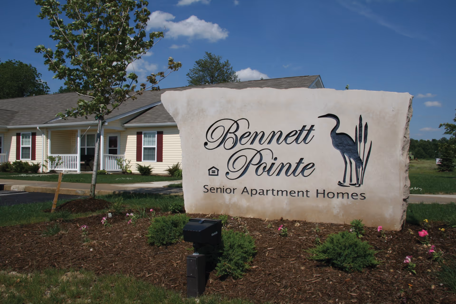 Large stone sign with the text 'Bennett Pointe Senior Apartment Homes' and a silhouette of a heron standing among cattails, set in a landscaped area with small bushes and flowers. In the background, there is a single-story building with beige siding, white trim, and maroon shutters under a blue sky with some clouds.