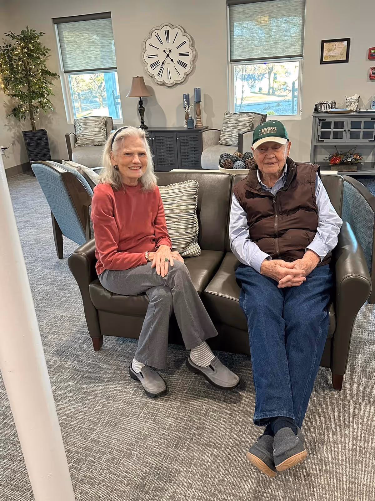 Two older adults seated on a couch in a well-lit retirement community common room with a large wall clock and windows behind them.