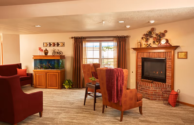 A cozy living room with two orange armchairs, one draped with a red blanket, facing a brick fireplace with a fire burning. There is a small wooden side table with a plant between the chairs. To the left, there is a wooden cabinet with a large fish tank on top and decorative items above it. The room has beige walls, a large window with brown curtains, and carpeted flooring.