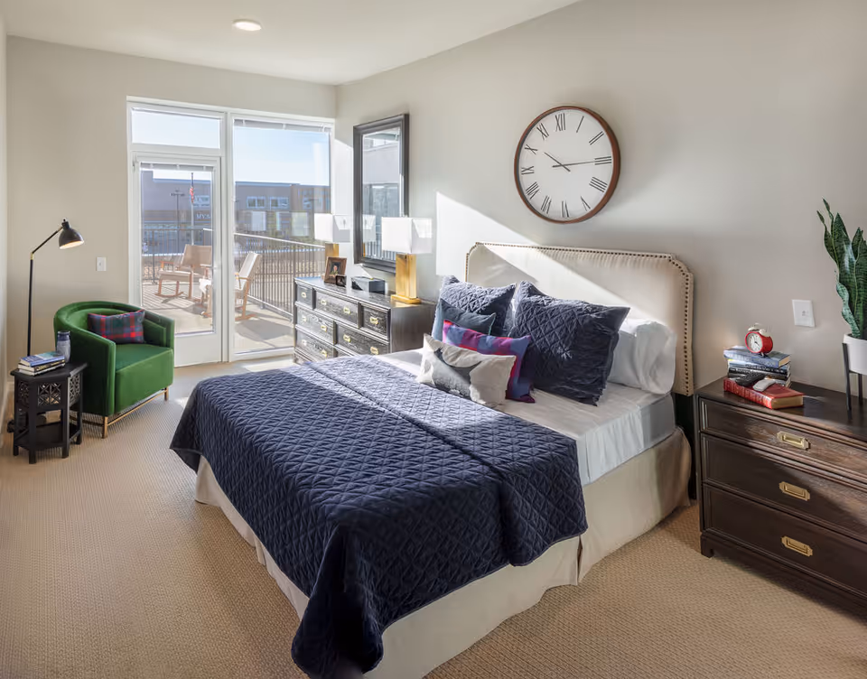 Sunlit modern bedroom with a navy quilted bed, bedside dressers, green armchair and sliding glass door opening to a balcony.