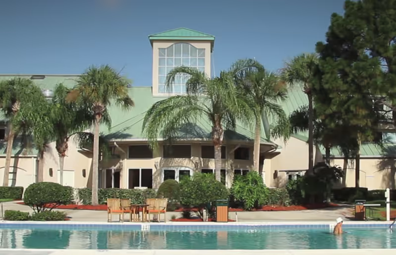 Exterior view of The Fountains of Melbourne facility featuring a building with a green roof, large windows, palm trees, and a swimming pool in the foreground with a person swimming.