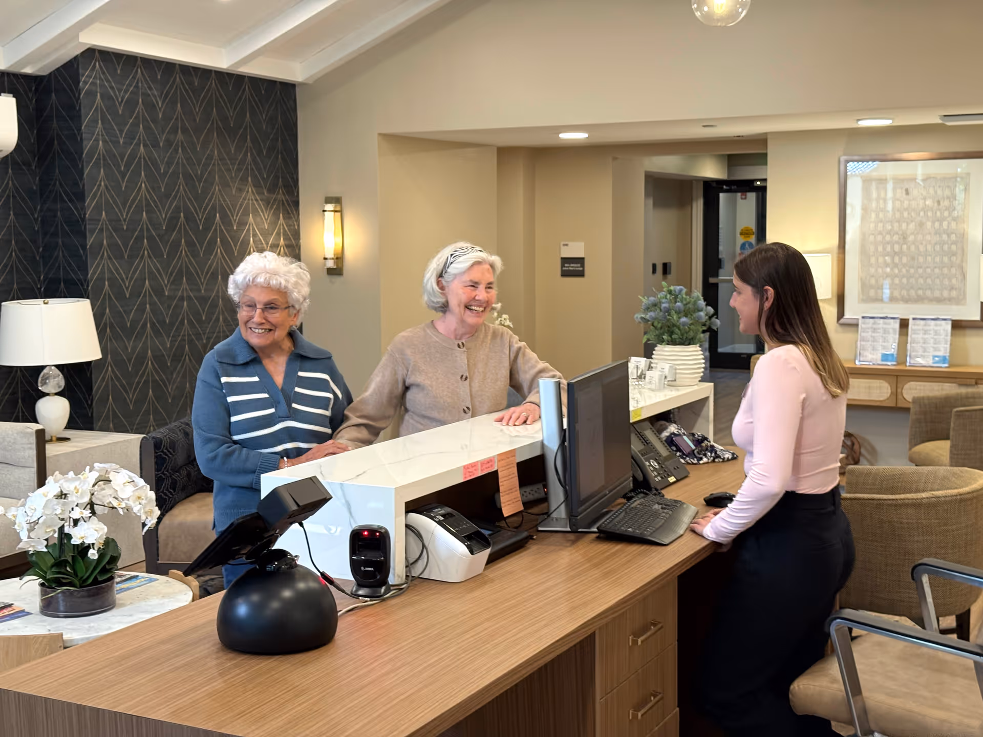 Two elderly women standing at a reception desk smiling and talking to a receptionist in a well-lit, modern interior space with comfortable seating and decorative plants.