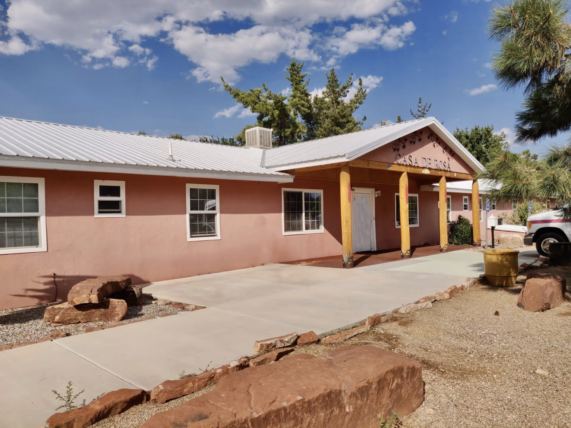 Exterior view of Casa de Rosa Assisted Living facility showing a single-story building with a light pink facade, white-framed windows, a metal roof, and a covered entrance supported by wooden pillars. The foreground includes a concrete walkway, rocks, and some trees under a partly cloudy blue sky.