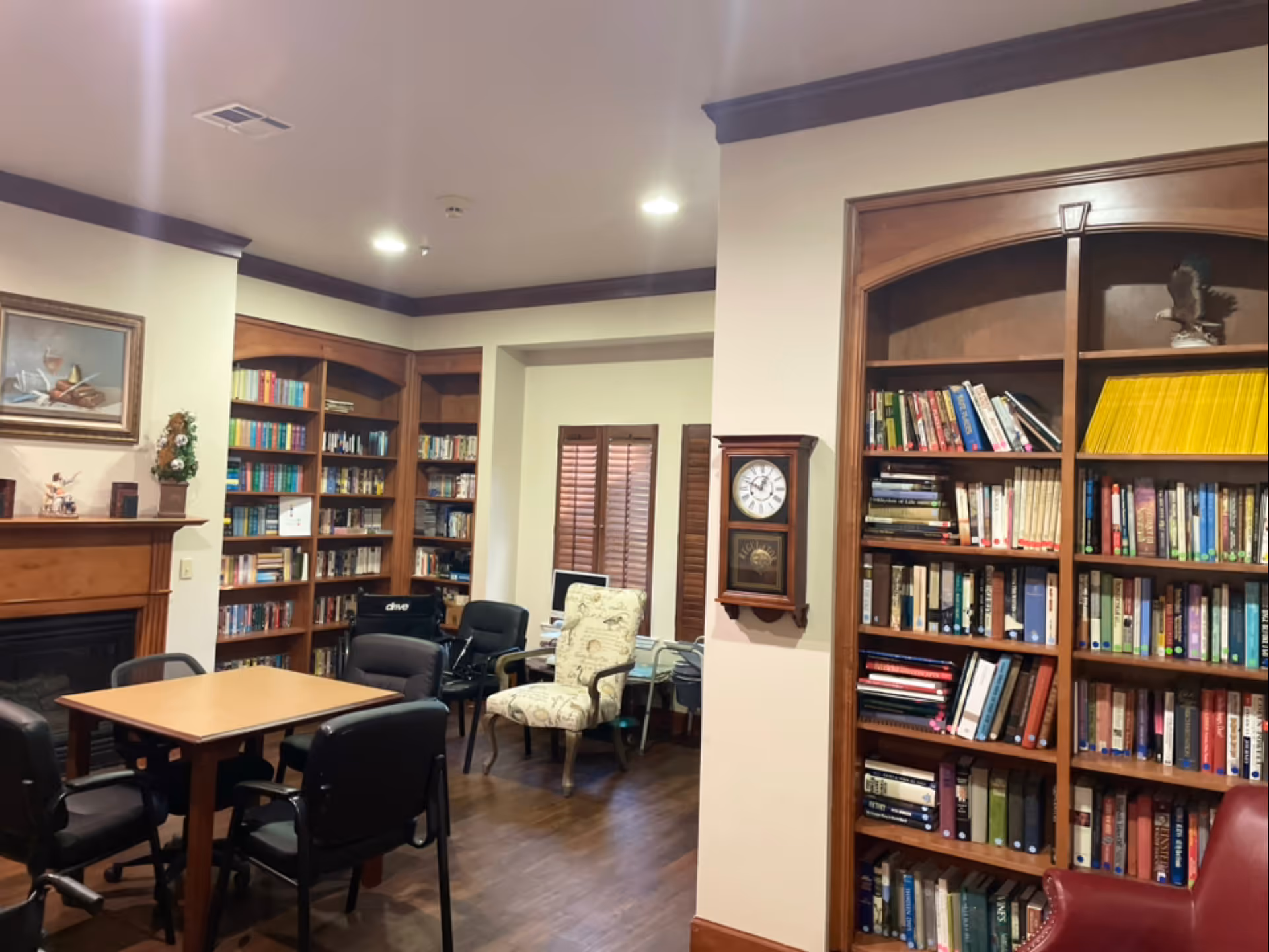 A cozy library or reading room with wooden bookshelves filled with books, a wooden table surrounded by black chairs, a fireplace with a mantel decorated with small items, a wall clock, and a comfortable armchair near a window with wooden shutters.