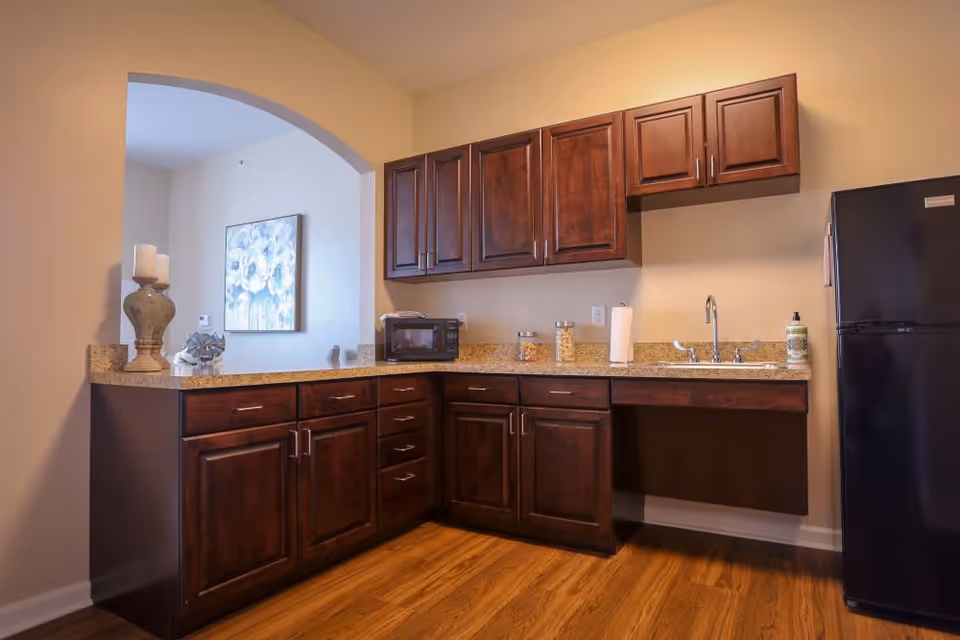A kitchen area with dark wooden cabinets, a granite countertop, a black refrigerator, a microwave, a sink with a faucet, and decorative items including a large candle holder and a piece of artwork visible through an arched opening.