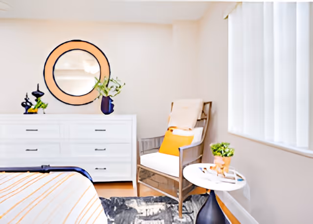 A bright bedroom featuring a white dresser with a round mirror above it, decorative items and a plant on top. There is a wooden armchair with white cushions and an orange pillow next to a small round table holding a plant and some books. The room has a window with vertical blinds and a bed with striped bedding partially visible in the foreground.