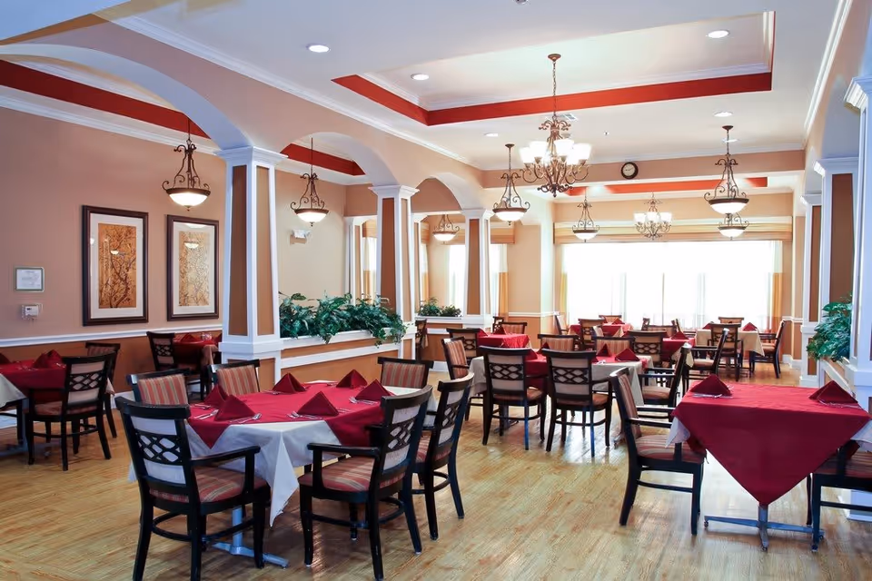 A spacious dining room with multiple tables covered in white and red tablecloths, each set with folded red napkins. The room features wooden chairs with cushioned seats, beige walls with white trim, decorative arches, hanging chandeliers, framed artwork, and large windows allowing natural light to fill the space.