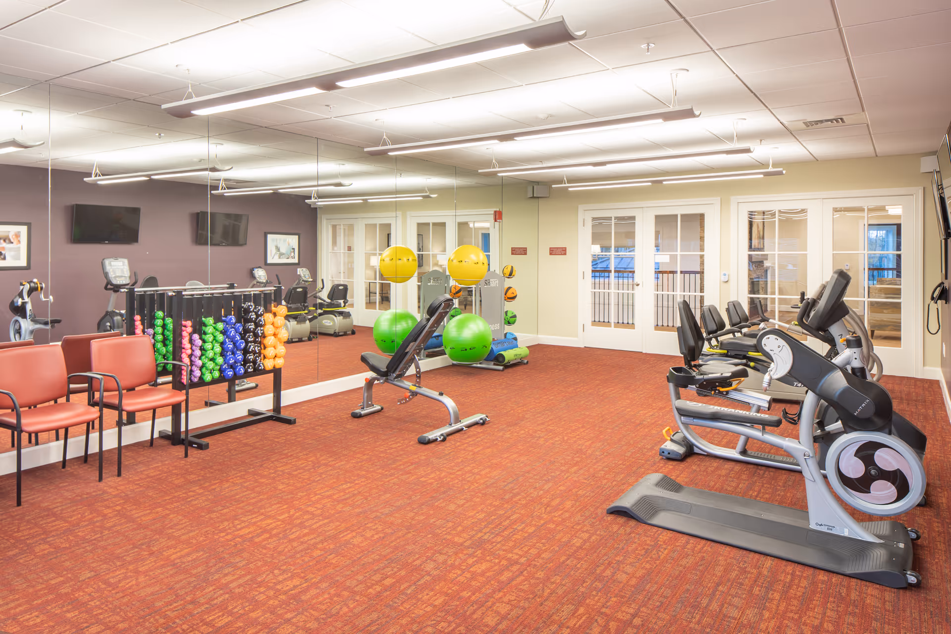 Bright fitness room with cardio machines, weight rack, exercise balls, mirrored wall and seating.