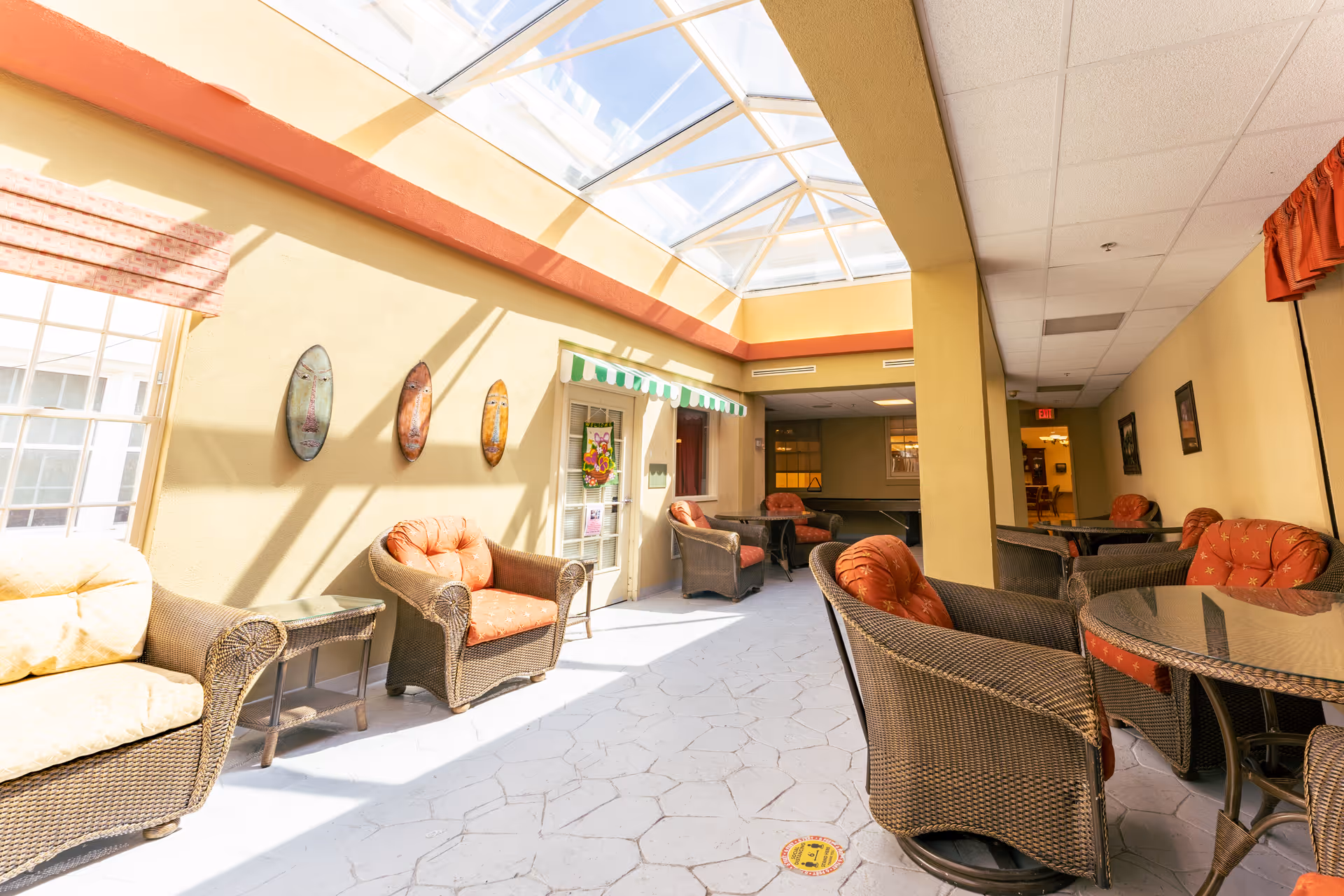 A bright indoor seating area with wicker chairs and cushioned seats arranged around glass-top tables. The space has a high ceiling with large skylights allowing natural light to flood in. The walls are painted yellow with decorative masks hanging on one side and a door with a green and white striped awning above it. The floor has a stone tile pattern.