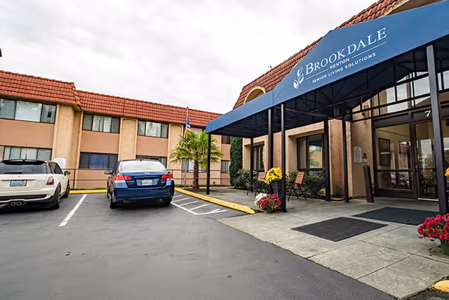 Entrance of a senior living building with a blue canopy reading Brookdale, a paved parking area and two parked cars.