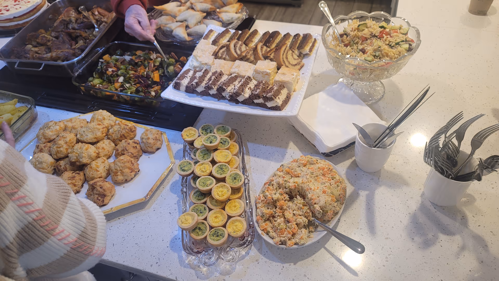 A buffet-style spread of various foods on a white countertop including biscuits, mini quiches, a bowl of salad, a plate of assorted desserts, roasted vegetables, chicken wings, and a bowl of a mixed salad dish. Utensils and napkins are arranged nearby, with a person reaching for food.