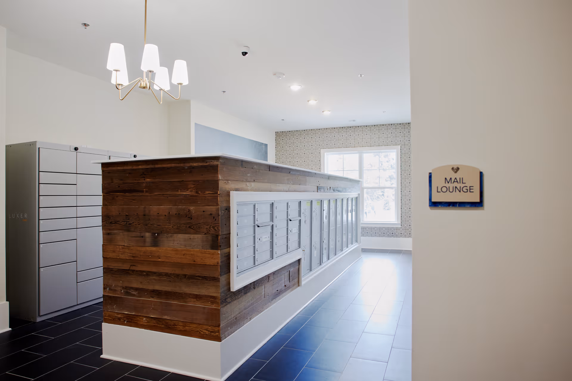 Interior view of a mail lounge area featuring a wooden and white mailbox unit with multiple mail slots, a gray parcel locker system on the left, a window letting in natural light, and a chandelier hanging from the ceiling.
