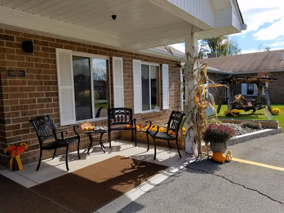 Covered front entrance of a brick senior living facility with metal chairs, seasonal flowers and pumpkins on the porch.