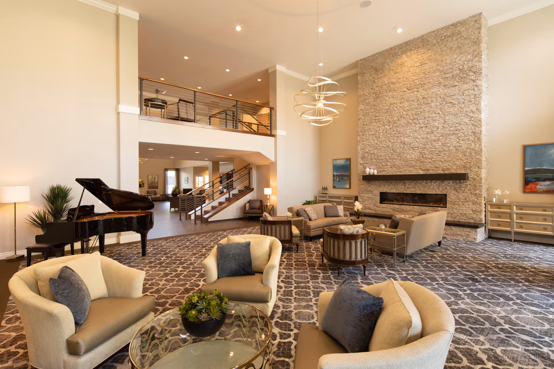 Spacious elegant communal living room with seating areas, a grand piano, a tall stone fireplace, and a mezzanine overlooking the space.