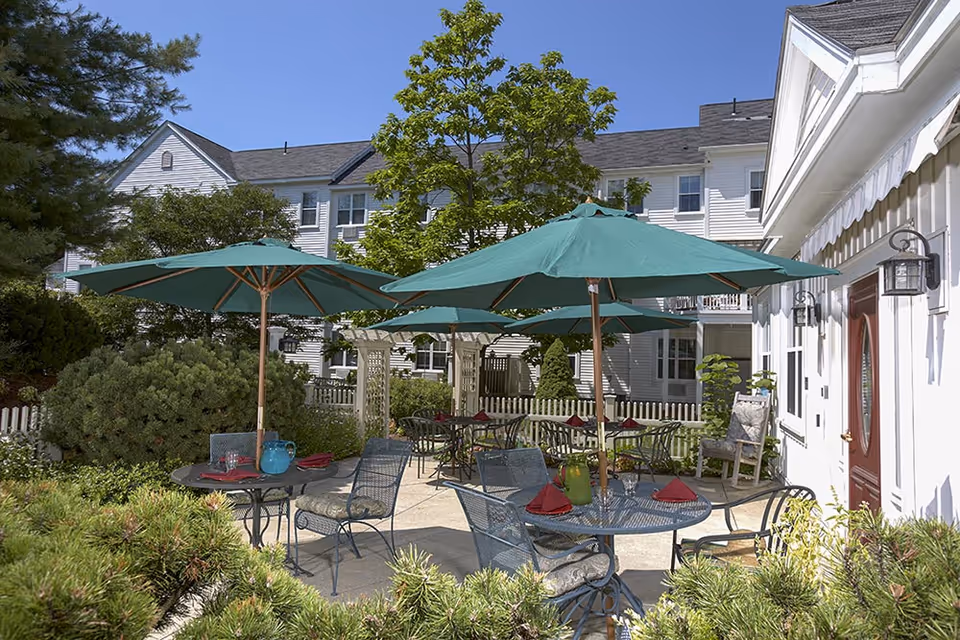 Courtyard patio with round tables, green umbrellas and metal chairs beside a white multi-story building and landscaped greenery.