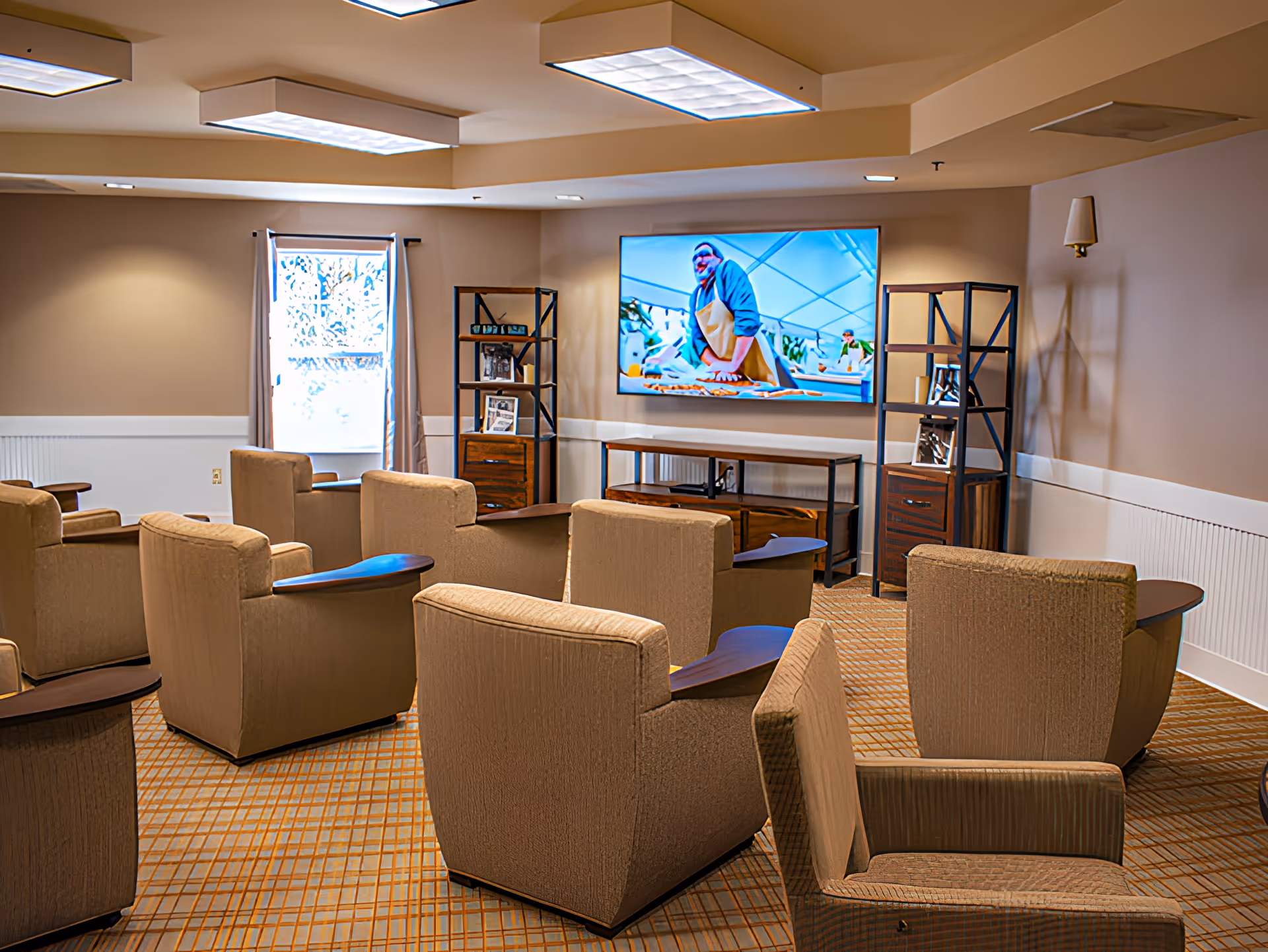 A cozy common room with several beige armchairs arranged in rows facing a large flat-screen TV mounted on the wall. The room has a patterned carpet, two shelving units on either side of the TV, a window with curtains, and soft overhead lighting.