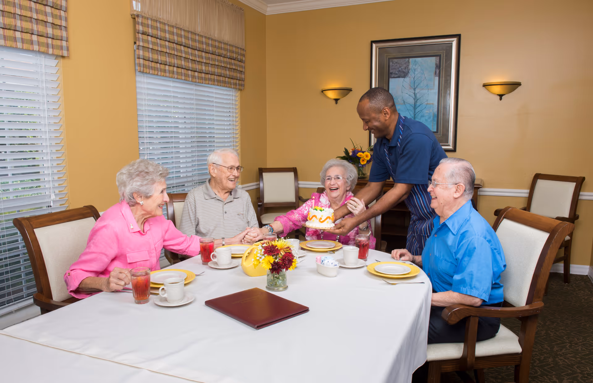 A caregiver serves a small cake to smiling elderly residents seated around a dining table in a dining room.