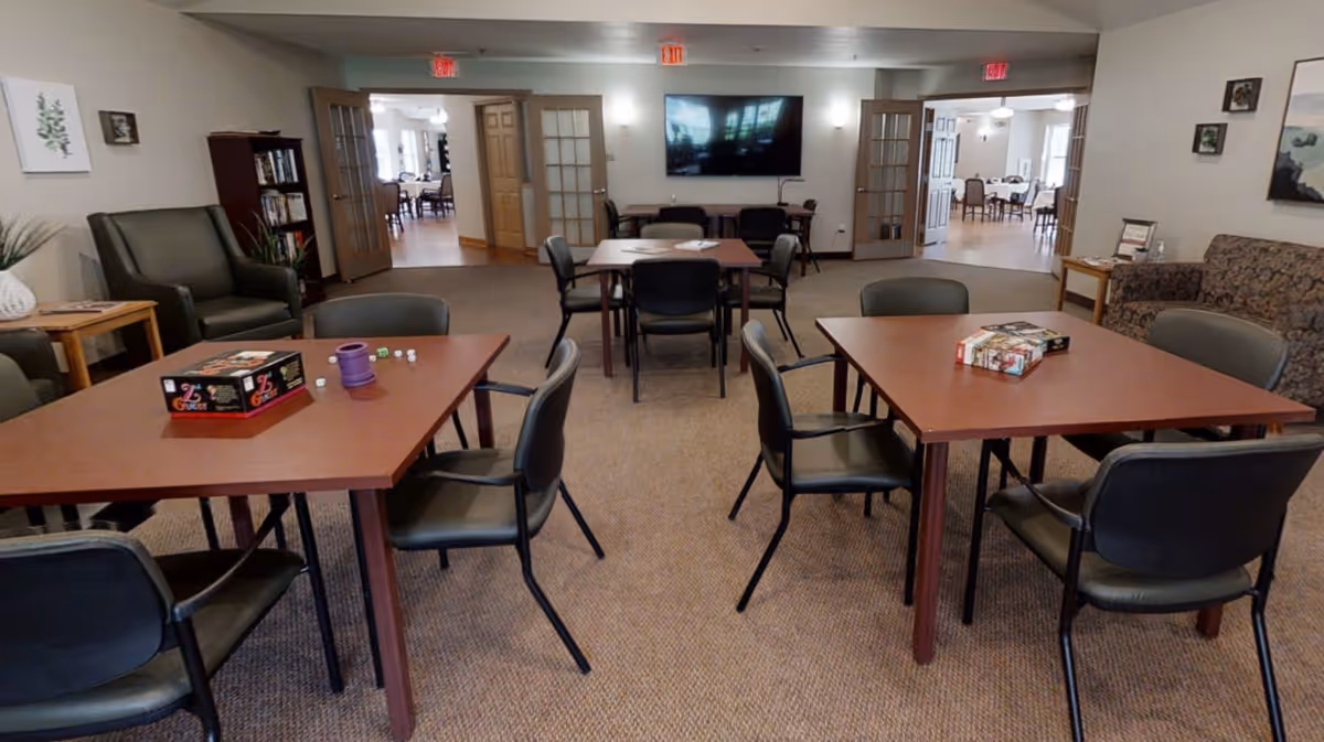 A common area in a senior living facility with several square tables surrounded by chairs. On the tables are board games and game pieces. The room has a carpeted floor, a large flat-screen TV mounted on the wall, and comfortable seating including armchairs and a sofa. There are double doors leading to another room with more tables and chairs.