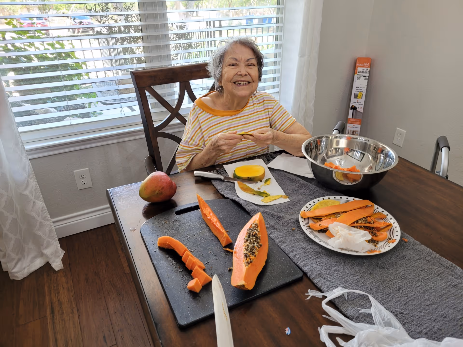 An elderly woman sitting at a wooden dining table in a bright room with large windows and white curtains. She is smiling and peeling a piece of fruit. On the table are a cutting board with sliced papaya, a whole mango, a large metal bowl, a plate with fruit peels, and a knife. The floor is wooden, and there is a walker next to the table.