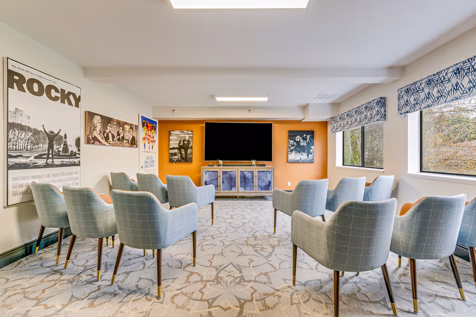 Small screening room with rows of light-blue upholstered chairs facing a wall-mounted TV and movie posters on the walls.