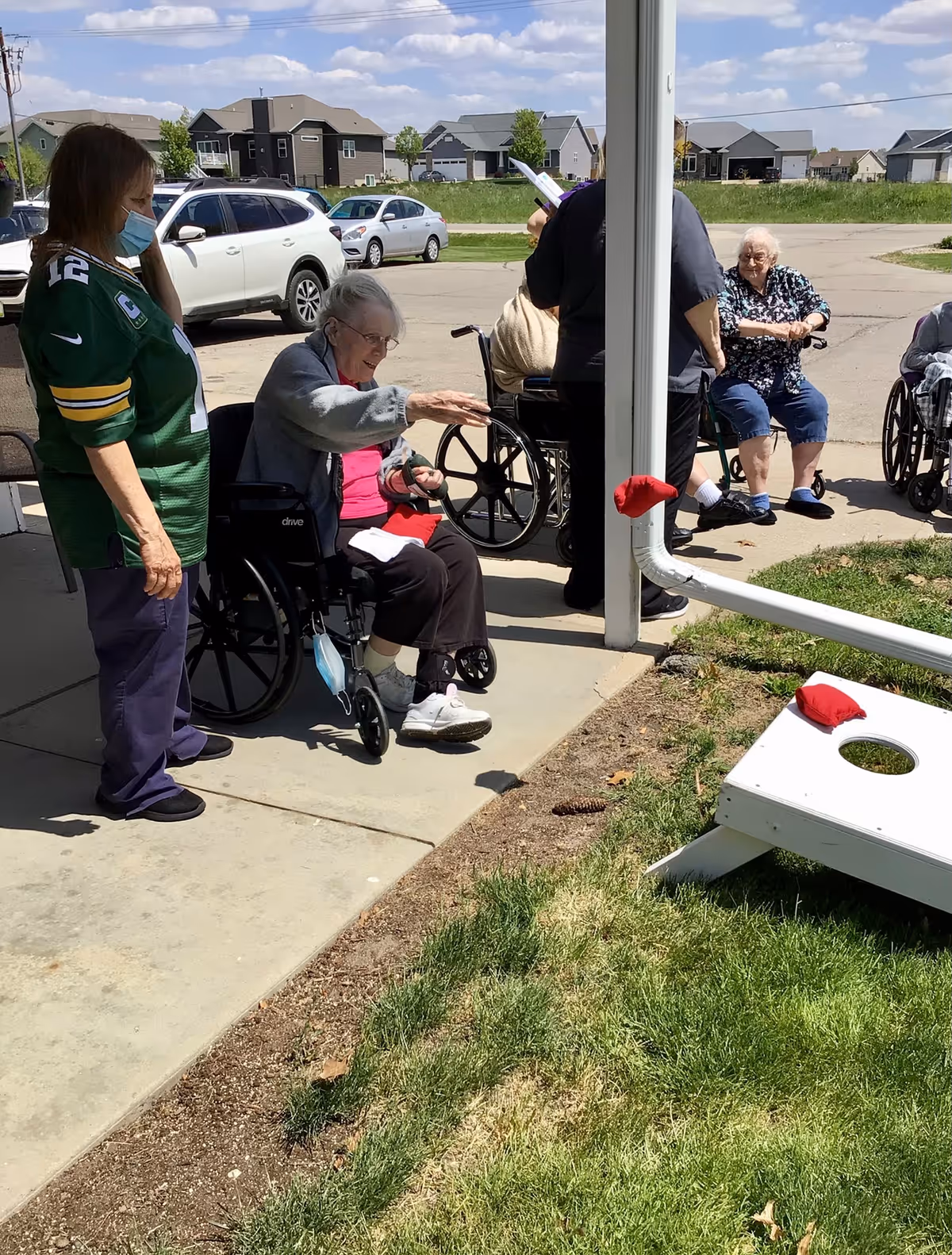 Several elderly individuals, some in wheelchairs, are gathered outside on a sunny day near a cornhole game set. A woman wearing a green sports jersey and a face mask stands beside an elderly woman in a wheelchair who is reaching out towards the cornhole board. Other elderly people are seated or standing nearby, with houses and parked cars visible in the background.